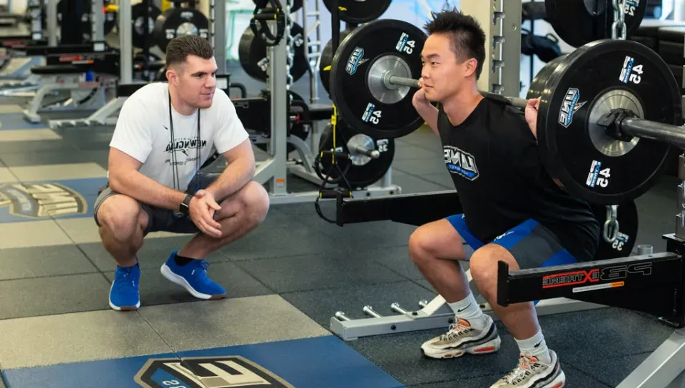 A student coaches another student performing squats with a barbell on their back